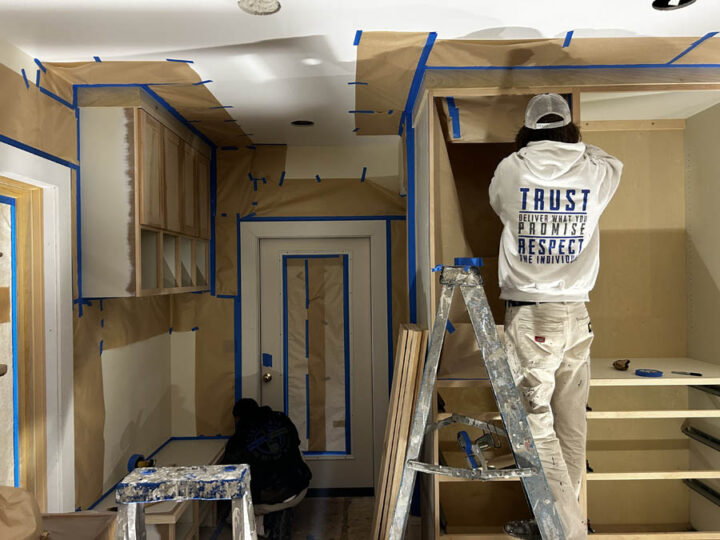 Two painters from Pearl Painters in Portland prepare kitchen cabinets for painting. Masking tape lines the walls, cabinets, and door frames as one painter stands on a ladder while the other works below.