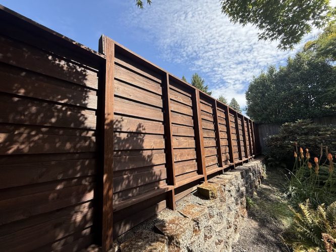 A tall wooden fence on a stone wall in Harvey's garden, greenery, and sky. Painted by Pearl Painters, Portland.