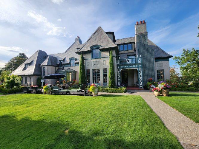 Painting by Pearl Painters shows a large house with peaks and chimneys, green lawn, flower pots, and furniture in Portland.