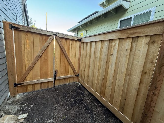 Wooden fence with gate between two houses, bare soil below, sunlight left. Pearl Painters project in Portland, July 2025.