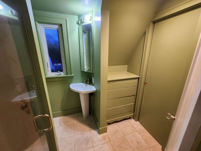 A small green bathroom in Portland with pedestal sink, mirror, window, built-in dresser, tiled floor, and glass shower door painted by Pearl Painters.