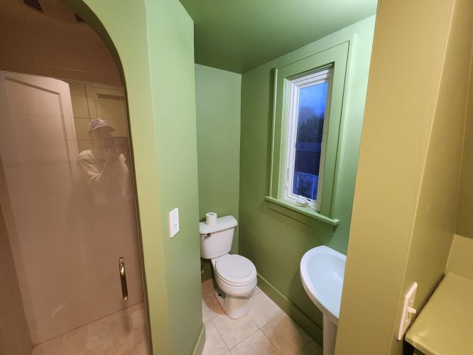In April 2025, a painter from Pearl Painters in Portland is partially visible in the glass shower door of a small green interior bathroom with a toilet, sink, and window.