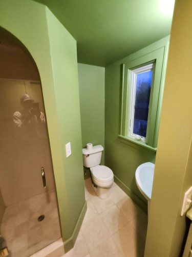 Painted by Duc in Portland, this small green bathroom features a toilet with a roll of toilet paper on the tank, a shower with glass door, a window, and a compact sink.