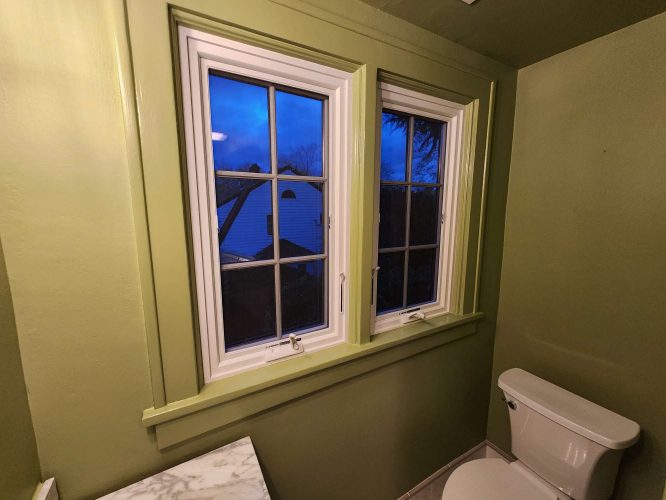 A painter from Pearl Painters in Portland showcases an interior bathroom with green walls, a white toilet, and two white-framed windows overlooking a house and tree at dusk. This painting could inspire April 2025 renovations.