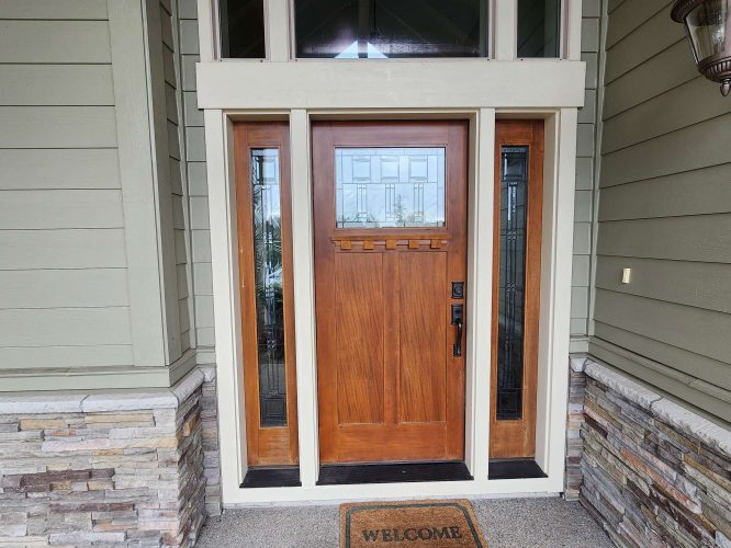 A Hansen exterior wooden front door with glass panels, set in green siding and stone accents, features a "WELCOME" doormat. Painting by Pearl Painters, Portland.