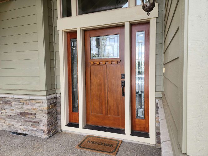 Portland painters from Pearl Painters recommend a Hansen wooden front door with glass panels, a "WELCOME" doormat, and stone-accented siding for an updated entryway this April 2025.