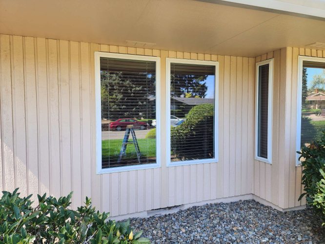 Photo of a beige house exterior in Portland, taken by Ferlitsch in April 2025. Includes two large windows reflecting a ladder, cars, and trees. Shrubs line the outside above a gravel area. Suitable for showcasing painting work by Pearl Painters or another local painter.