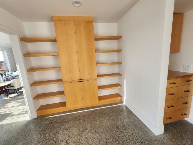 Painted built-in wooden shelving unit from the Isadore collection, featuring a central cabinet and open shelves, located in a room with polished concrete floors and white walls. Available April 2025. Pearl Painters, Portland.