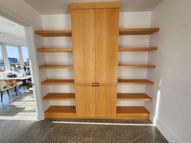 Isadore Cabinets offers a light wood built-in shelving unit with six open shelves per side and a central two-door cabinet, set against a white wall. Available in Portland April 2025. For painting, contact Pearl Painters.
