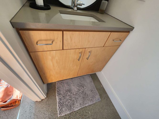 The Isadore wooden bathroom vanity, located in Portland, has a gray countertop, white sink, two cabinet doors, and two drawers. A patterned bath mat sits on polished concrete. Ideal for an updated look in April 2025. Painting by Pearl Painters.