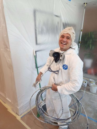 A painter from Pearl Painters in Portland stands by cabinets prepared for painting, wearing protective clothing and holding a paint sprayer, ready for an April 2025 renovation by Isadore.