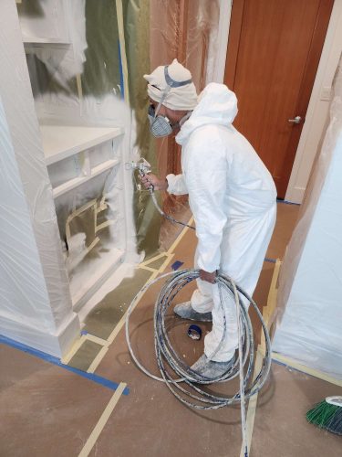 A painter from Pearl Painters in Portland uses a spray gun to paint built-in cabinets, with nearby surfaces protected by plastic and tape. Scheduled for completion by Isadore in April 2025.