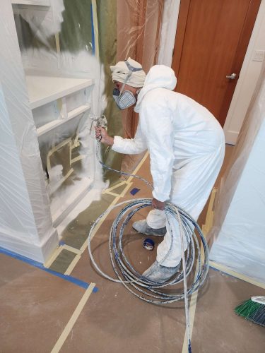 In April 2025, a painter in protective gear from Pearl Painters uses a sprayer to paint built-in cabinets in Portland; floors and surroundings are protected with plastic and tape.