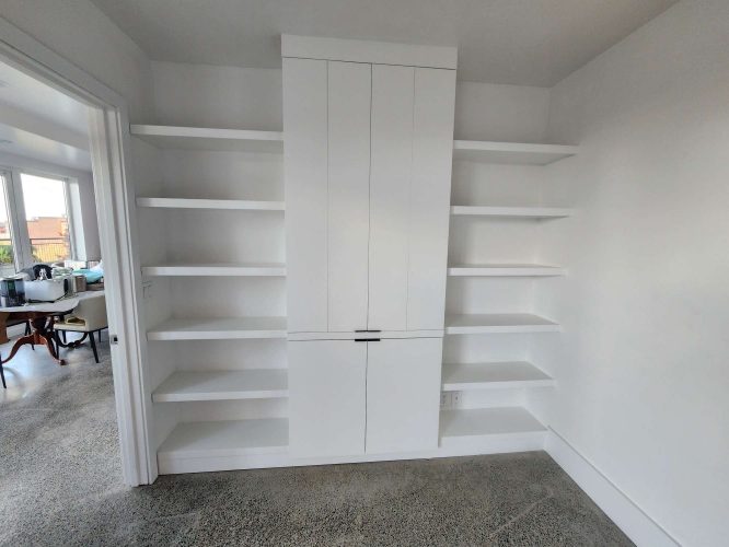 White built-in shelving unit by Isadore, with central cabinets and open shelves, against a white wall and concrete floor. Available April 2025 in Portland. Painted by Pearl Painters.
