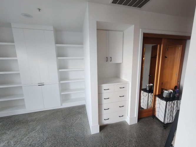 A modern room in Portland features built-in white cabinets, shelves, drawers, a polished concrete floor, and a cart with toiletries near a wooden sliding door—prepared for Isadore's arrival in April 2025. Painting by Pearl Painters.