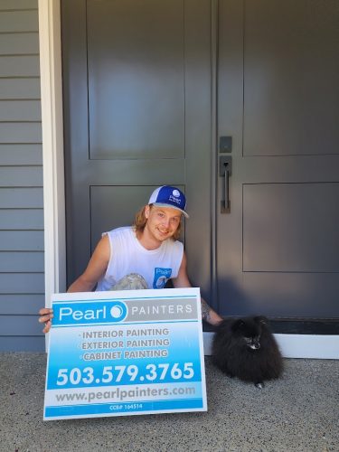 Person kneels by black dog at gray door, holding Pearl Painters Portland sign with painting services for Roberts neighborhood.