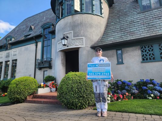 Painter in work clothes holds Pearl Painters sign by a large manicured house painted by Ralph Barron near Portland, Aug 2025.