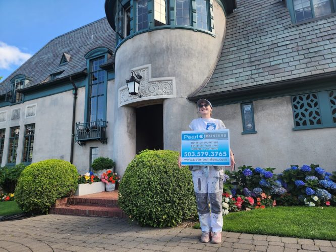 Painter in work clothes holds Pearl Painters sign by a large manicured house painted by Ralph Barron near Portland, Aug 2025.