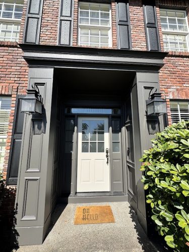 Brick house front entrance with gray door, black trim; painted by Pearl Painters, Portland. Shrubs and "OH HELLO" doormat visible.
