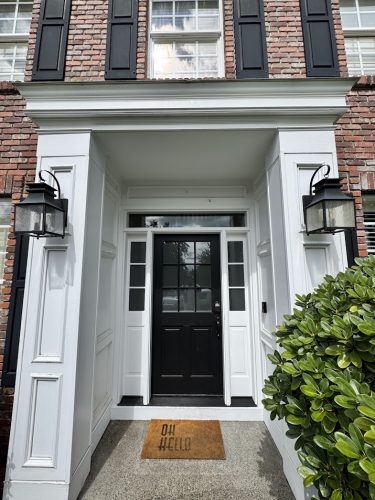 Black front door with glass panes, white trim, brick walls; lantern lights, "OH HELLO" mat. Painted by Pearl Painters, Portland.