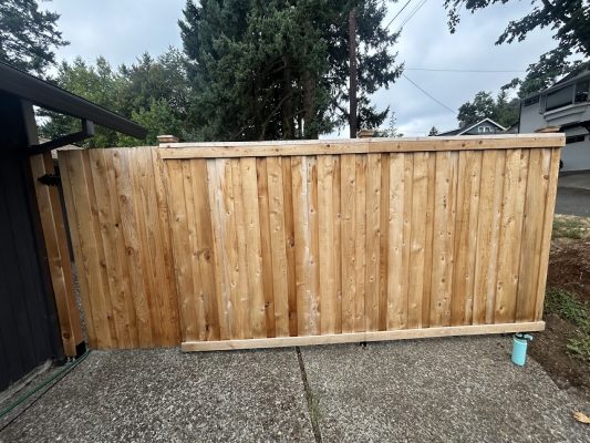 A wooden fence by a concrete driveway with trees and houses; blue water bottle at right, likely left by a Pearl Painters painter in Portland.
