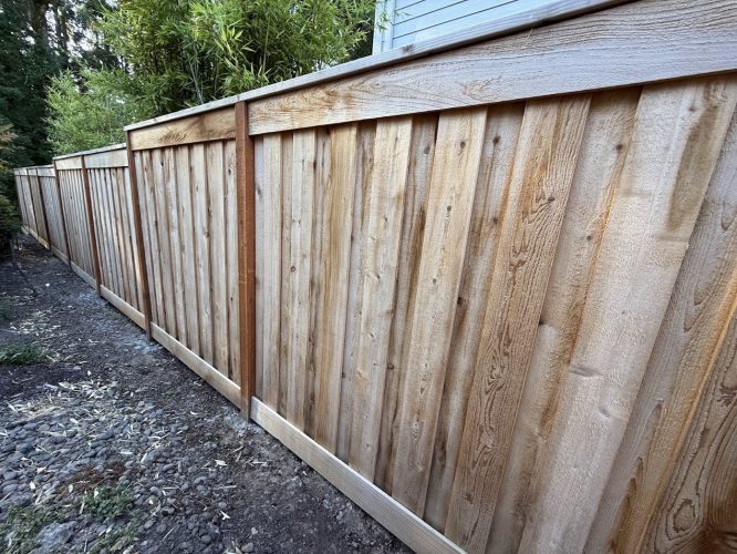 Long new wooden fence by dirt path, painted by Pearl Painters, Portland. Trees and building visible. Photo: Smith, July 2025.