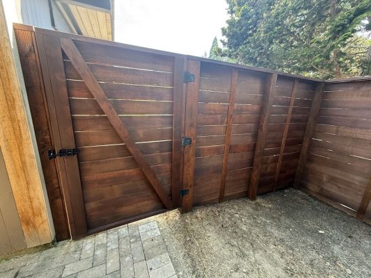 A Portland painter inspects a wooden fence and gate with horizontal slats in Harvey, July 2025, for Pearl Painters.