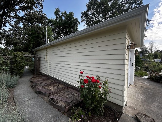 Pearl Painters painting a beige-sided garage with concrete path and garden beds, red rose bush, under partly cloudy July sky in Portland.