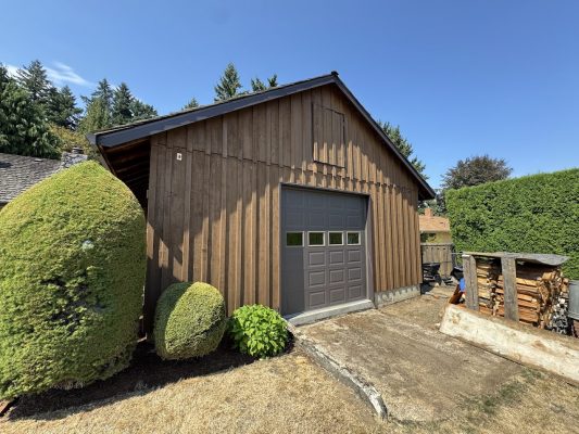 Craytor wooden shed with gray garage door, bushes, firewood, and driveway painted by Pearl Painters in Portland, August 2025.