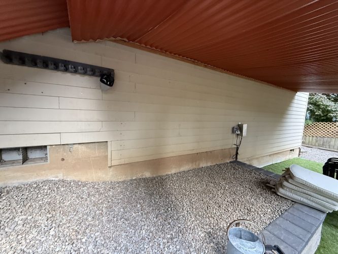 Exterior gravel area next to beige building with utility boxes and red roof at Montoya, seen during Pearl Painters’ work in Aug 2025.