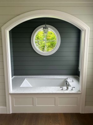 Portland painter Duffy styled this bathroom for May 2025, featuring a white bathtub in a marble alcove, round window, chandelier, and dark green shiplap walls. Pearl Painters handled the painting.