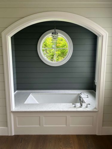Portland painter Duffy styled this bathroom for May 2025, featuring a white bathtub in a marble alcove, round window, chandelier, and dark green shiplap walls. Pearl Painters handled the painting.