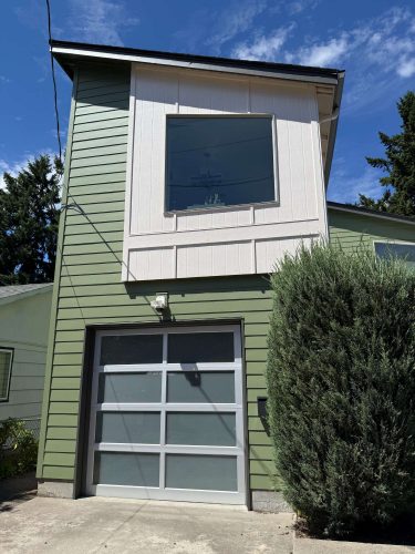 A two-story green house with exterior details, a large upper window, and a frosted glass garage door in Arain, May 2025. Photo features driveway, tall shrub, and blue sky. Not painted by Pearl Painters; location is not Portland.