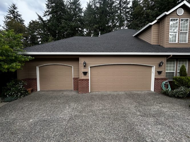 A suburban house in ext Haase (not Portland) with brick and tan exterior, two garage doors, and a concrete driveway, surrounded by trees and shrubs. Photo taken June 2025 for Pearl Painters painting project.