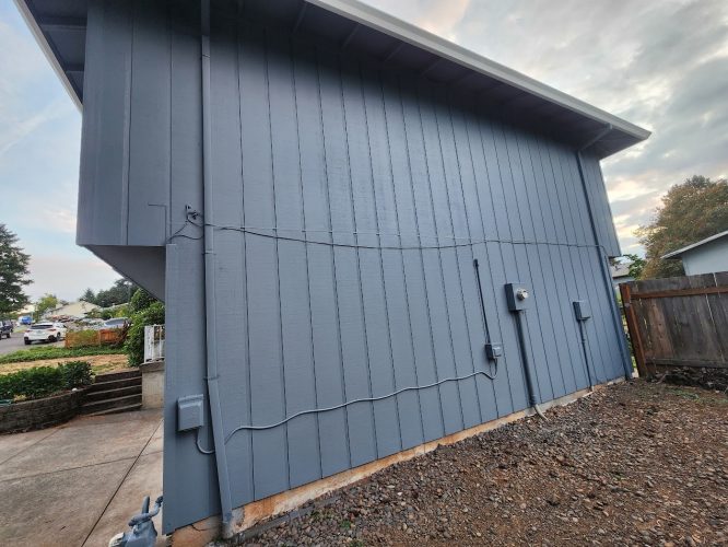 Side view of the gray Mahoney exterior house with vertical siding, utility boxes, wires, and rocky ground next to a wooden fence in Portland, June 2025. Painting by Pearl Painters.