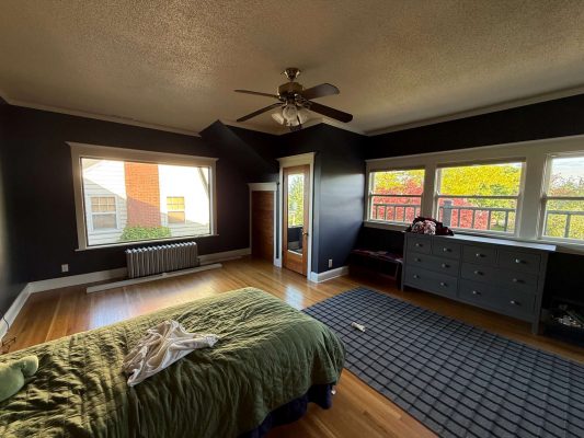This photo, taken in May 2025, shows a painted bedroom in Portland with dark walls, a green bedspread, gray dresser, ceiling fan, large windows, and balcony access. Pearl Painters was the painter for this project.