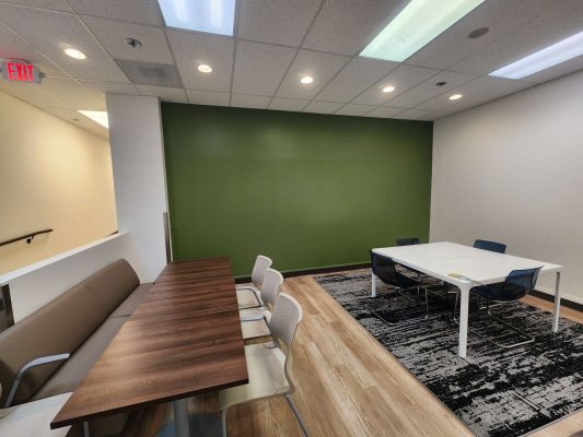 The modern Jones office in Portland has a green accent wall, painted by Pearl Painters, with a wooden table and chairs on the left and a white meeting table on a patterned rug to the right. Photo from May 2025.