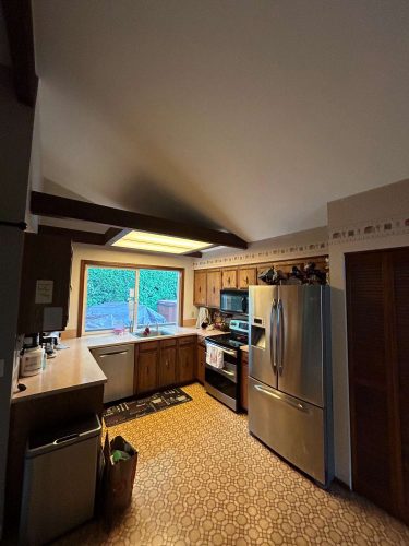 A kitchen in Portland features wood cabinets, stainless steel appliances, patterned linoleum flooring, and a large window overlooking greenery. Painted by Pearl Painters.