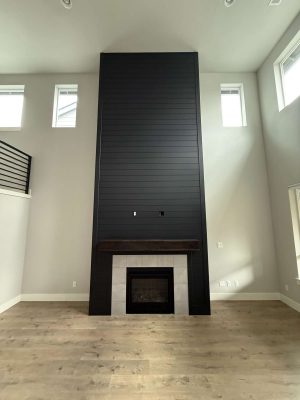 A Pearl Painters painting project in Portland: a modern living room with a black vertical shiplap accent wall, fireplace, wood mantle, light wood floors, and four high windows, completed May 2025.