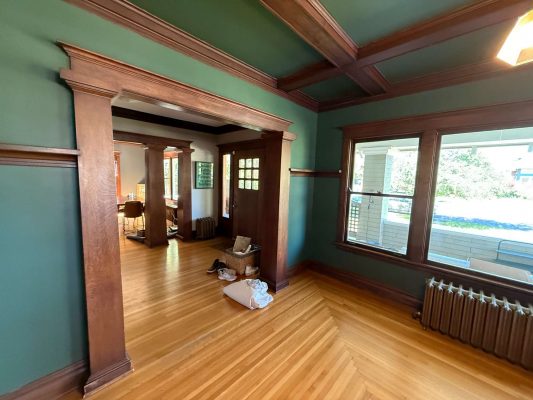 A room in Portland painted green with wood trim and floors, featuring a large window, radiator, and open doorway; bags and items are on the floor near the doorway. Painter: Pearl Painters.