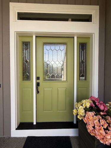A green exterior front door with decorative glass panels, white trim, and a black handle is shown with a planter of pink and yellow flowers beside it; painted by Pearl Painters in Portland.