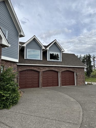 A house in Portland with brick and gray siding, three brown garage doors, two upper windows under gabled roofs, and a wide concrete driveway is scheduled for exterior painting by Pearl Painters in June 2025.