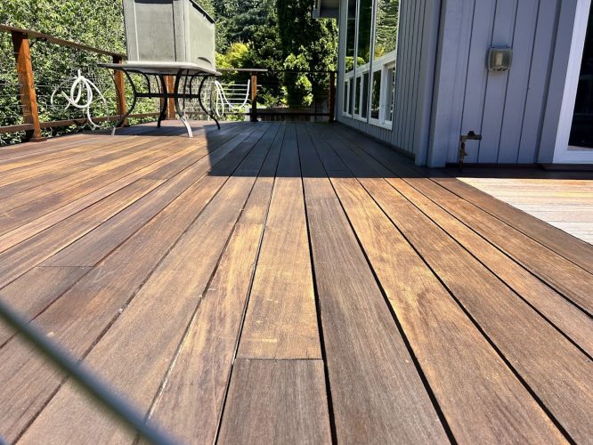 Wooden deck painted by Shainsky, bordered by a railing, next to a house with large windows and outdoor furniture in Portland. Painted for gatherings in June 2025 amid trees and greenery. Pearl Painters provided painting services.