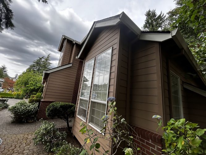 A painter from Pearl Painters in Portland captured a brown house with large windows and red brick accents amid shrubs and trees under a cloudy June 2025 sky.