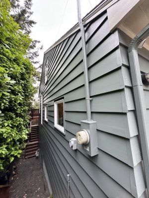 Gray-sided house in Carlton with utility meter, conduit pipes, narrow walkway, small windows, and dense greenery. Photo taken June 2025 by Pearl Painters.