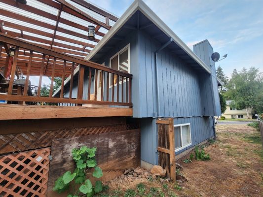 A blue house with a raised wooden deck and lattice skirting is painted in the ext Mahoney neighborhood. The painting was done by Pearl Painters, a Portland painter, June 2025. A small fenced area and nearby plant indicate summer greenery.