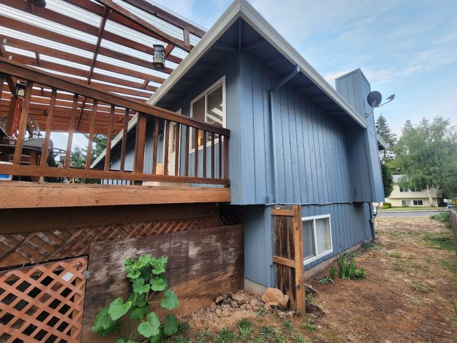 A blue house with a raised wooden deck and lattice skirting is painted in the ext Mahoney neighborhood. The painting was done by Pearl Painters, a Portland painter, June 2025. A small fenced area and nearby plant indicate summer greenery.