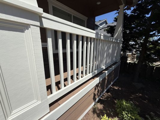 White wooden porch railing with brown trim, part of an Emory house, with greenery and trees in the background under a clear June 2025 sky. Painted by Pearl Painters, Portland painters.