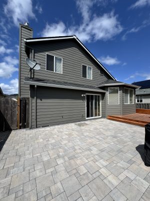 Photo of a two-story gray house with a satellite dish, sliding glass door, and multiple windows, next to a large patio and wooden deck. Taken in May 2025 near Horn. Possible project location for Pearl Painters, Portland painting company.