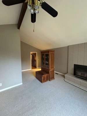 A cozy living room painted by Pearl Painters in Oneal, Portland, features beige carpet, gray walls, a wooden cabinet and chest, brick fireplace, ceiling fan with lights, and a vaulted ceiling with exposed beam.